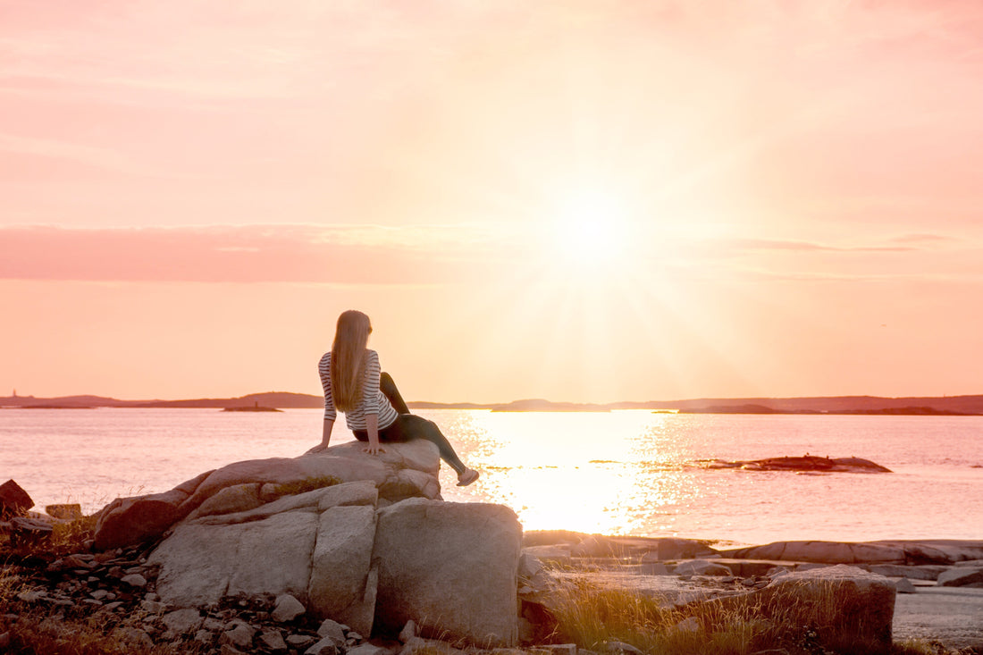 Summer skincare blog cover woman sitting on a rock watching golden glow of sun setting.