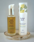Two skincare products, Nordic Renewal Elixir Facial Oil and Twilight Birch Nighttime Serum, on a wooden stand with a gray background