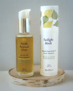 Two skincare products, Nordic Renewal Elixir Facial Oil and Twilight Birch Nighttime Serum, on a wooden stand with a gray background