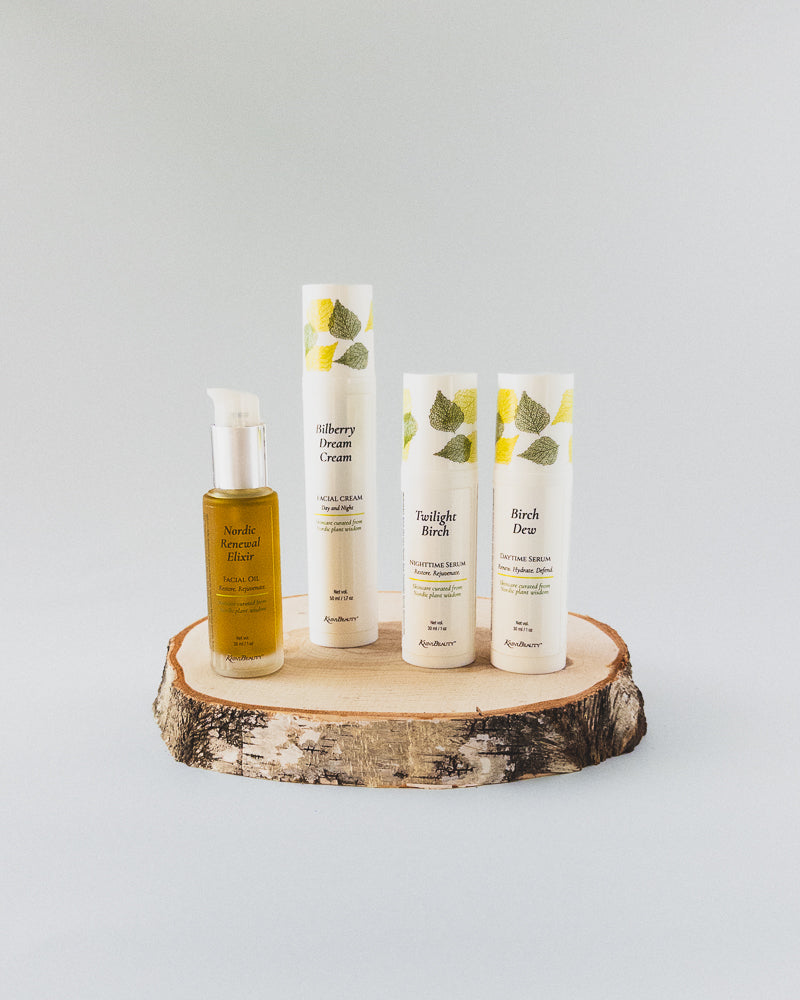 Four skincare products from Kasvi Beauty Birch Collection arranged on a rustic wood slice pedestal against a light gray background. The set includes a 'Nordic Renewal Elixir Facial Oil' bottle and 'Bilberry Dream Cream Facial Moisturizer', 'Birch Dew Daytime Face Serum', 'Twilight Birch Nighttime Face Serum' white airless bottles  packaging decorated with green and yellow birch graphics.