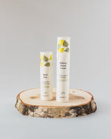 Two white cosmetic airless pump bottles labeled 'Bilberry Dream Cream Face Moisturizer' and 'Birch Dew Daytime Face Serum'  by Kasvi Beauty, decorated with green and yellow leaves, displayed on a rustic birch wood slice against a light gray background.