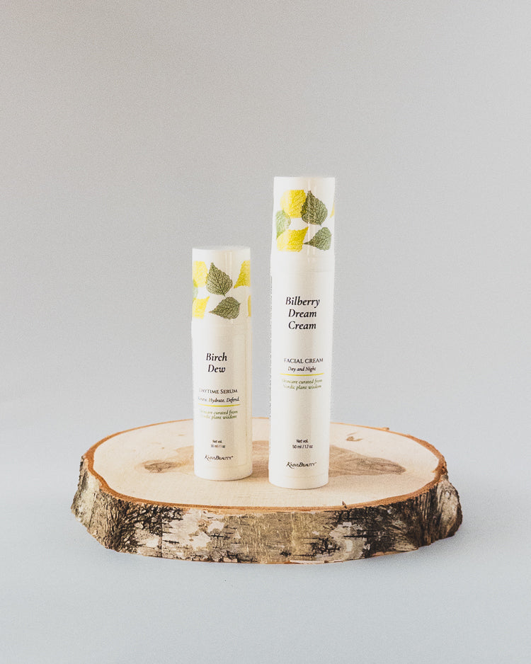 Two white cosmetic airless pump bottles labeled 'Bilberry Dream Cream Face Moisturizer' and 'Birch Dew Daytime Face Serum'  by Kasvi Beauty, decorated with green and yellow leaves, displayed on a rustic birch wood slice against a light gray background.