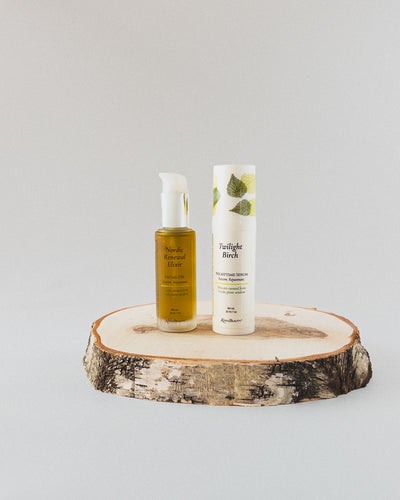 Two skincare products from the brand Nordic Renewal Elixir Facial Oil with a white pump, and a white airless pump of Twilight Birch Nighttime Renewal Serum with green leaf graphics, displayed on a round birch wood slice against a light gray background.